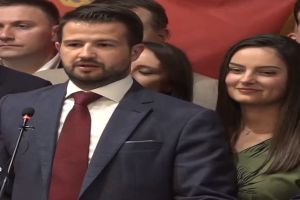 Ein Mann im Anzug und Krawatte steht an einem Podium mit einem Mikrofon und spricht zu einer Menge mit einem Plakat im Hintergrund, wahrscheinlich auf eine Regierungssperre für soziale Medien reagierend.