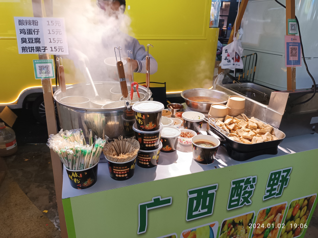 Ein Mann steht vor einem chinesischen Streetfood-Truck und bereitet Gerichte mit Schüsseln, Tassen, Löffeln, Zangen und anderen Utensilien auf dem Tresen zu, während im Hintergrund ein Fahrzeug geparkt ist.