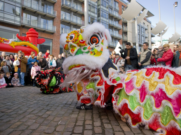 Ein farbenfrohes chinesisches Neujahrsfest in Amsterdam mit einem Löwen tanzen vor einer Zuschauermenge, darunter einige mit Kameras, vor Buildings und einem klaren blauen Himmel.