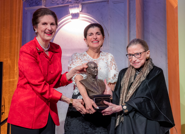 Drei Frauen stehen zusammen, wobei die mittlere Frau eine Statue hält, vor einer Wand mit einem Licht, einschließlich Ruth Bader Ginsburg und Nancy Pelosi, die mit einem Lifetime Achievement Award geehrt werden.