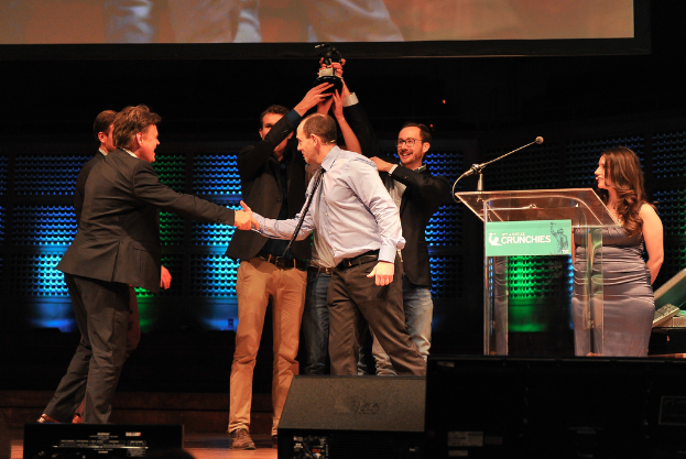 Zwei Männer in schwarzen Anzügen heben einen Preis hoch, während ein Mann links die Hand schüttelt und eine Frau rechts steht.