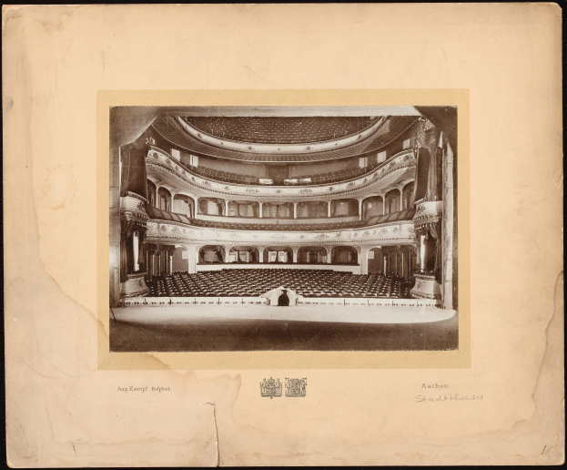 Altes Schwarz-Weiß-Foto des Auditoriums der Royal Albert Hall in London, das die Bühne, Stuhlreihen, Säulen, Geländer und Text mit einem Logo unten auf einem dunklen Hintergrund zeigt.