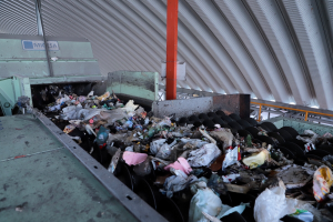 Ein großer Müllhaufen auf einem Förderband in einer Recyclinganlage, mit Maschinen im Vordergrund und einem Metalldach darüber.