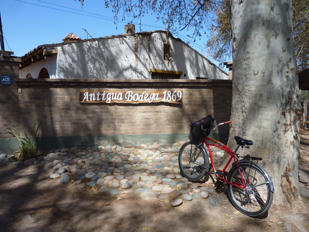 Ein rotes Fahrrad steht auf einem großen Baumstamm mit Steinen auf dem Boden, mit einer Begrenzungsmauer, auf der "Antike Bodega 1869" steht und einem weißen Haus im Hintergrund.