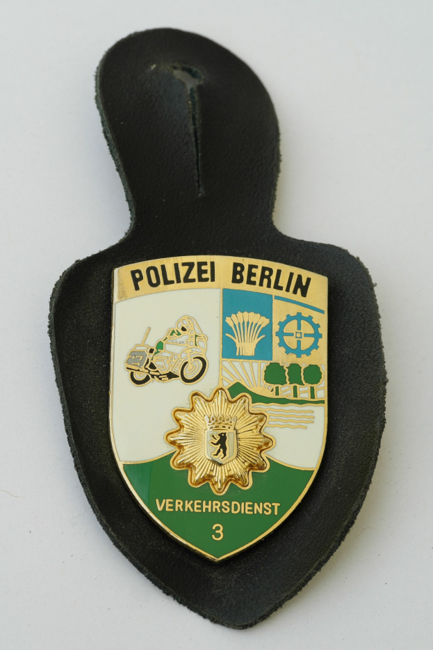 Ein Polizeischild mit der Aufschrift 'Polizei Berlin' darauf, das ein blaues Schild mit einem weißen Stern in der Mitte zeigt, umgeben von einem weißen Rand, auf einer Oberfläche platziert.