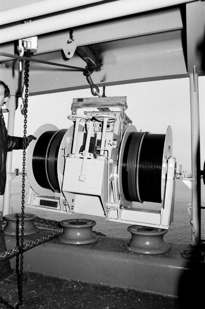 Ein Mann in Schuhen steht neben einer großen Maschine mit einer Kette in einem zeitlosen Schwarz-Weiß-Bild.