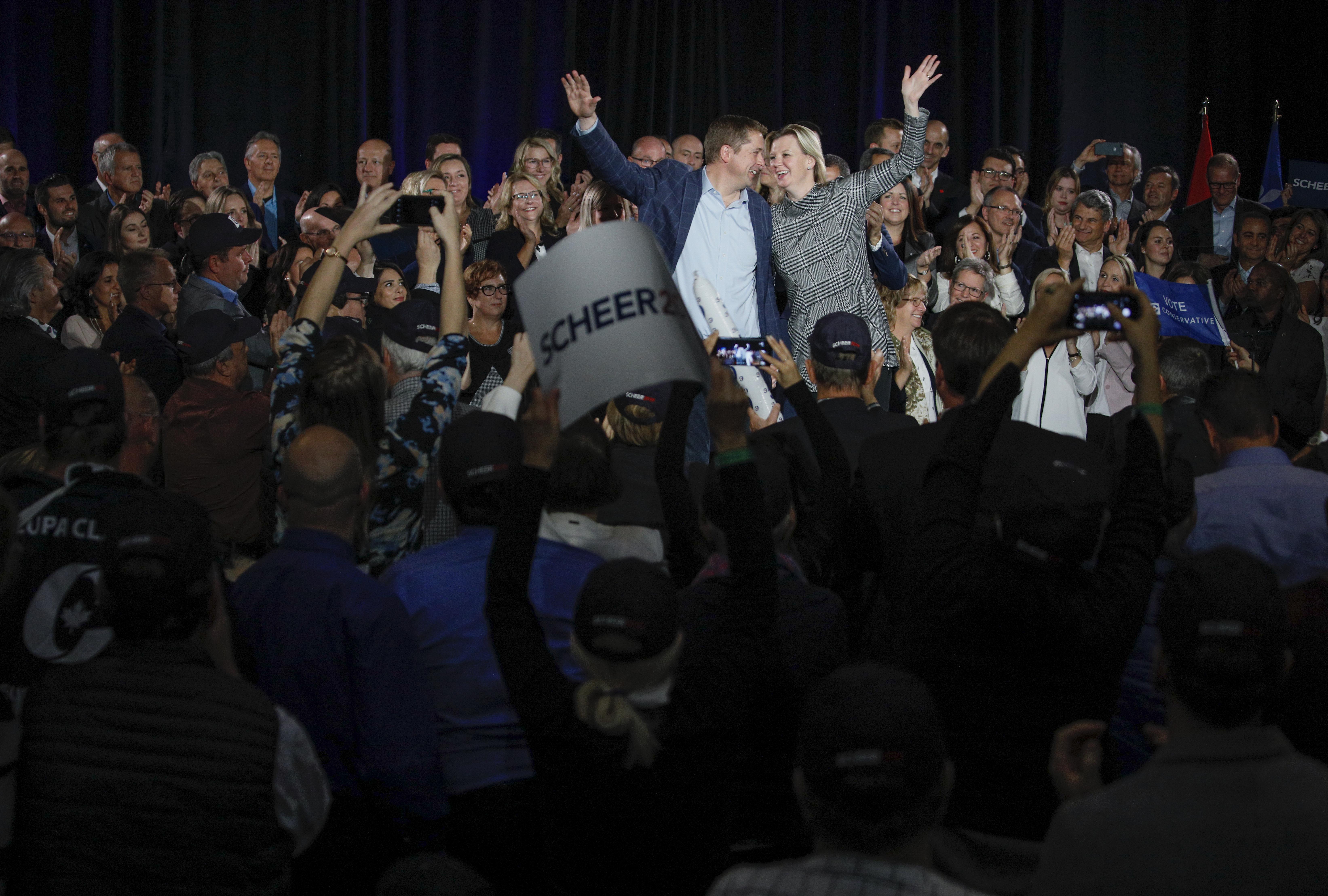 Ein Mann und eine Frau stehen vor einer Menge, mit einigen, die Schilder und Mobiltelefone halten, und Fahnen und einem Vorhang im Hintergrund.