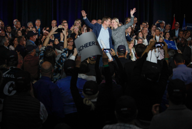 Ein Mann und eine Frau stehen vor einer Menge, mit einigen, die Schilder und Mobiltelefone halten, und Fahnen und einem Vorhang im Hintergrund.