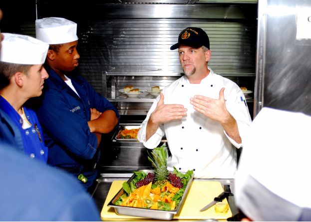 Ein Mann in Kochuniform spricht mit einer Gruppe von Menschen in einer Küche, mit einem Tablett mit Essen, einem Messer und anderen Gegenständen auf dem Tisch davor und Tabletts mit Essen im Hintergrund.