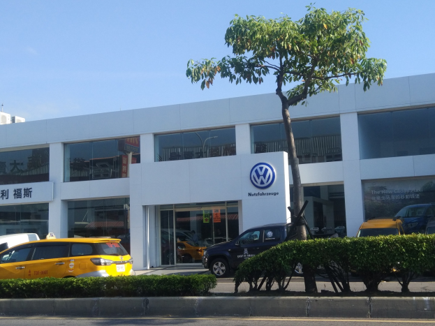 VW-Händler mit Glaswänden und -fenstern, Autos davor geparkt, umgeben von Pflanzen und einem Baum.