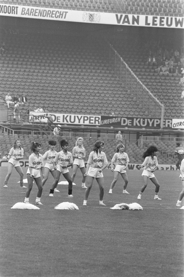 Schwarzes Bild von Frauen auf einem Fußballfeld mit Zuschauern auf den Rängen und Bannern im Hintergrund.