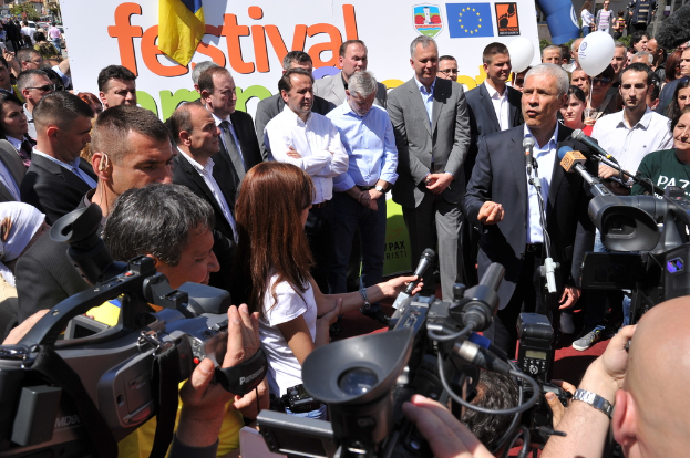 Eine Gruppe von Menschen steht auf einer Straße und hält Kameras und Mikrofone in den Händen, mit einem Plakat, Bäumen, Fahrzeugen, Topfpflanzen, einem Ballon, Pfosten und Gebäuden im Hintergrund.