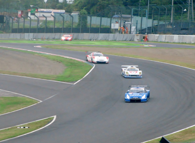 Eine Gruppe von Autos, die auf einer Strecke fahren, umgeben von Gras, einem Maschendrahtzaun, Pfosten, Bäumen und einem klaren blauen Himmel im Hintergrund.