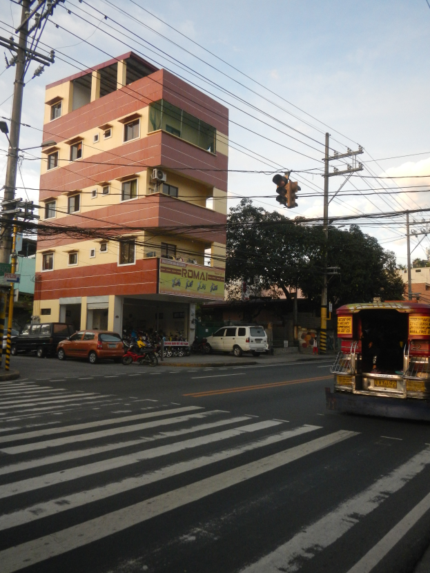 Eine belebte Stadtstraße mit Fahrzeugen, darunter ein Tuk-Tuk im Vordergrund, Strommasten, eine Ampel, Gebäude mit Fenstern, Bäume und ein bewölkter Himmel.