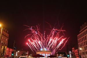 Eine belebte Straßenkreuzung in Berlin an Silvester, voller Menschen, Fahrzeuge und Gebäude, erleuchtet von Feuerwerk und Gebäudelichtern, die eine festliche Atmosphäre schaffen.