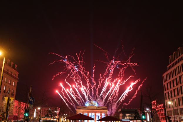 Eine belebte Straßenkreuzung in Berlin an Silvester, voller Menschen, Fahrzeuge und Gebäude, erleuchtet von Feuerwerk und Gebäudelichtern, die eine festliche Atmosphäre schaffen.