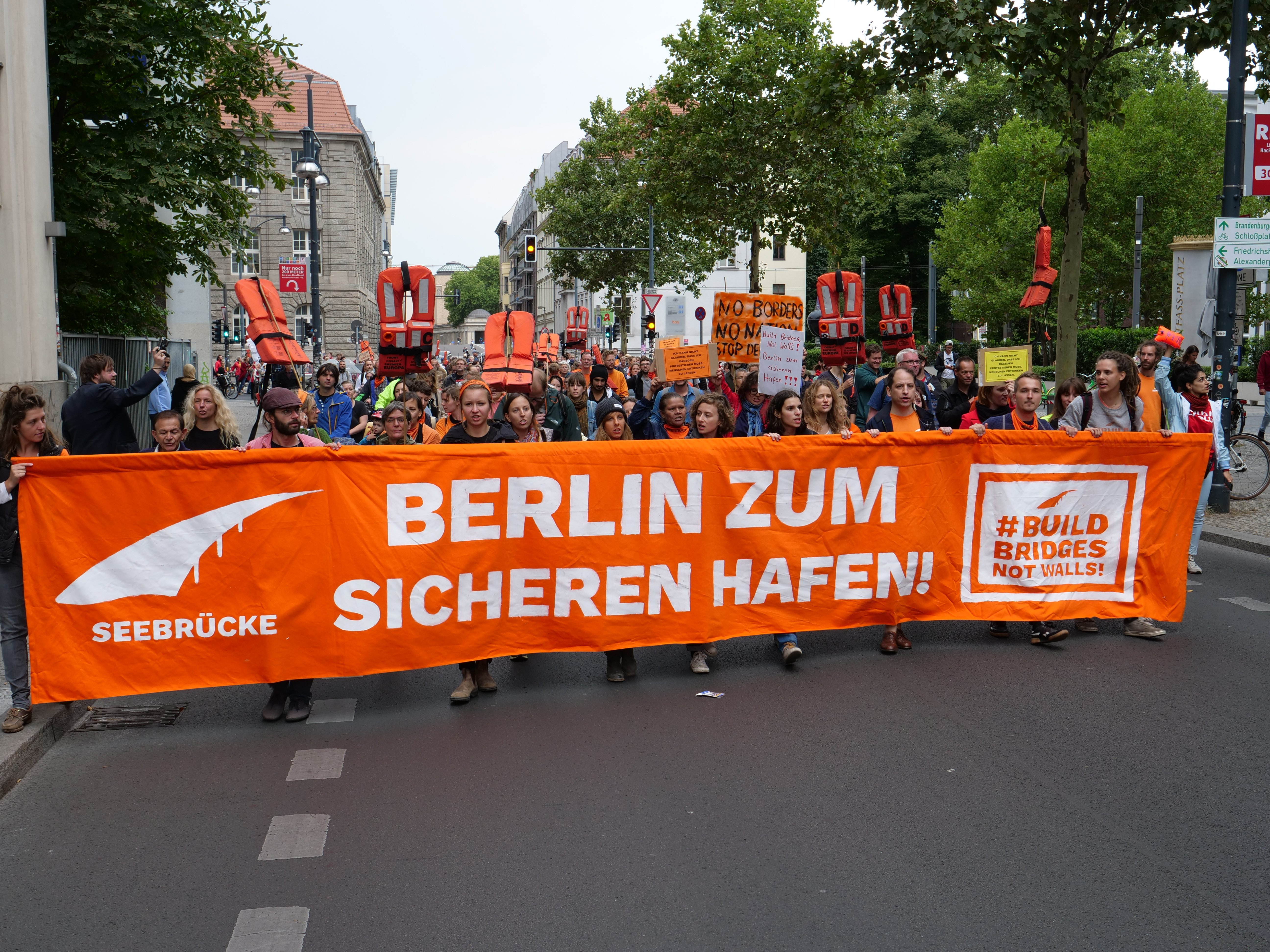Eine Gruppe von Menschen marschiert mit einem orangen Banner, auf dem 'Berlin Zum Sicherhen Hafen' steht, durch eine Straße. Im Hintergrund sind Fahrradfahrer, Bäume, Laternenpfähle und Gebäude unter einem klaren blauen Himmel zu sehen.