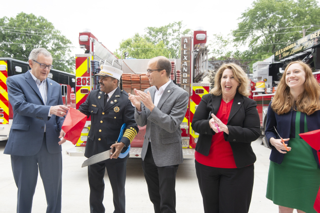 Gruppe von Menschen, die bei einer Einweihungszeremonie für die Alexandria Fire Department klatschen und lächeln, mit zwei Personen, die eine Schere und ein rotes Band halten, vor einem Feuerwehrauto, Bäumen, Drähten und einem klaren blauen Himmel im Hintergrund.