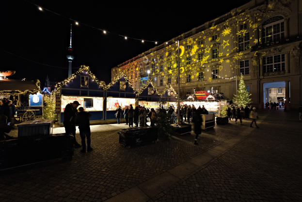 Ein lebendiger Weihnachtsmarkt in Berlin, Deutschland, mit Menschen um beleuchtete und geschmückte Stände, Gebäuden mit Fenstern im Hintergrund unter einem dunklen Himmel.