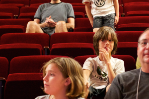 Ein Junge in einem weißen T-Shirt und schwarzen Hosen steht in der Mitte eines Kinosaals voller Menschen in roten Stühlen, umgeben von einer Atmosphäre der Vorfreude und Aufregung.