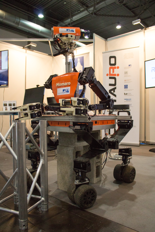 Ein vierrädriger Roboter sitzt auf einem Tisch in einem Raum, umgeben von Metallstangen, einem Fernseher auf einem Ständer, einer Fahne mit Text und einer Decke mit Lampen.
