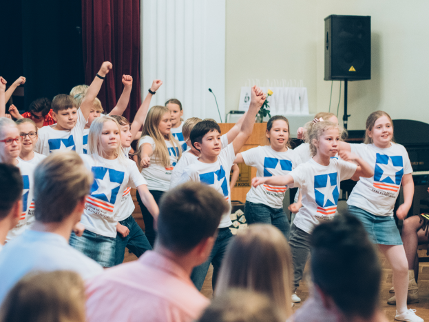 Eine Gruppe von Kindern in weißen Hemden und blauen Shorts tanzt auf einer Bühne vor einem sitzenden Publikum, mit einem Vorhang, Lautsprecher und einem Tisch mit einem Mikrofon im Hintergrund.
