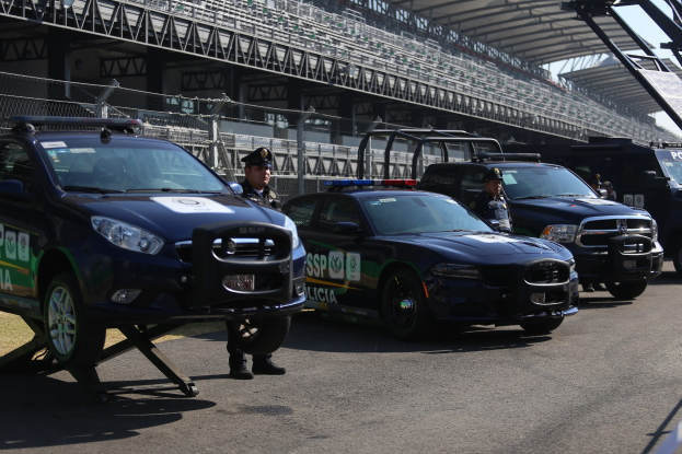 Polizeifahrzeuge vor einer Rennstrecke geparkt, Stadion mit Geländern und Maschendrahtzaun sowie Himmel im Hintergrund.