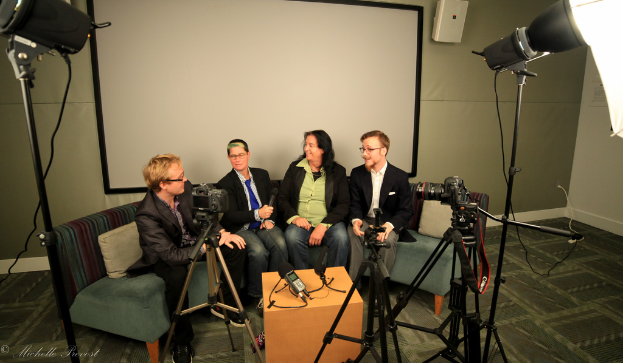Eine Gruppe von drei Personen sitzt auf einem Sofa und gibt ein Interview, wobei eine Person links in einem schwarzen Mantel und Hose, eine Person in der Mitte mit einem Mikrofon haltend und einem blauen Schlips sowie eine rechts lächelnde Person zu sehen sind. Ein Mikrofonrecorder befindet sich auf dem Tisch in der Mitte, Kameras sind auf der rechten und linken Seite sowie große Scheinwerferständer vorhanden, im Hintergrund ein Projektionsschirm.