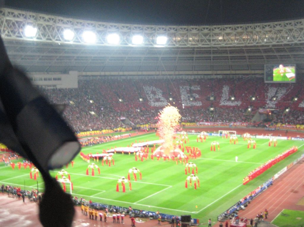 Ein Stadion mit Flutlicht, Eisenstäbe, Zuschauer, stehende Personen, Werbetafeln, Bildschirme, Überwachungskameras, Fahnen und Dekorationen.