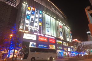 Ein Bus fährt auf einer Straße mit hohen Gebäuden, die mit Weihnachtsbeleuchtung geschmückt sind, vorbei an Straßenlaternen, Bäumen, Ampeln und verschiedenen städtischen Elementen unter einem sichtbaren Himmel.