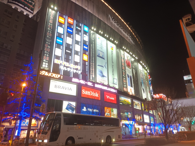 Ein Bus fährt auf einer Straße mit hohen Gebäuden, die mit Weihnachtsbeleuchtung geschmückt sind, vorbei an Straßenlaternen, Bäumen, Ampeln und verschiedenen städtischen Elementen unter einem sichtbaren Himmel.