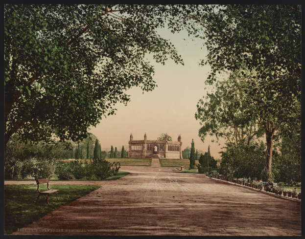 Ein altes Foto eines Parks mit einer Bank, umgeben von grünem Gras, Pflanzen und Bäumen, im Hintergrund ein Gebäude mit Säulen und Bögen und Text am unteren Bildrand.