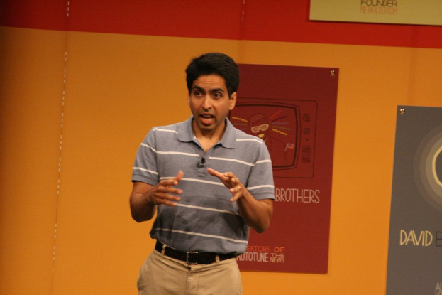 Ein Mann steht auf der Bühne und spricht mit einer Wand voller Plakate im Hintergrund.