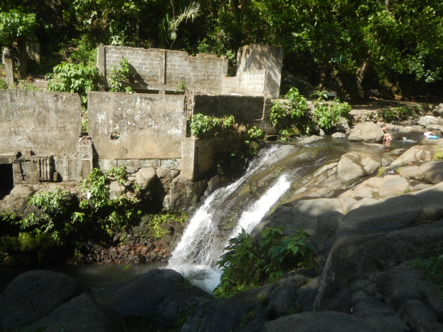 Ein kleiner Wasserfall ergäßt sich über eine steinige Klippe in einem grünen Dschungel, mit Menschen, die im Wasser schwimmen und einer Wand im Hintergrund.