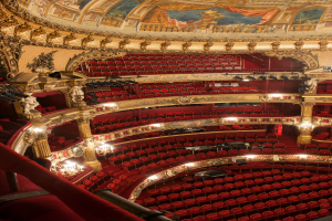 Ein Blick hinter die Kulissen des Opernhaus-Auditoriums in London, mit Sitzreihen, Geländern, Säulen, Lampen, Wandskulpturen und einer Deckenmalerei.