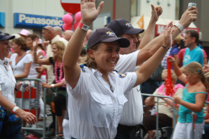 Eine Gruppe von Menschen steht vor einer Menge bei einem Umzug, einige tragen Mützen und halten Dosen, andere sitzen auf Stühlen, mit Geländern, Ballons und einem Gebäude mit Namensschildern im Hintergrund.