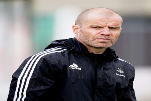 Ein kahlköpfiger Mann in einer schwarzen Jacke steht auf einem Fußballfeld, trägt eine marineblaue Jacke mit weißen Streifen und Kragen und hat die Hände gefaltet und einen ernsten Gesichtsausdruck.