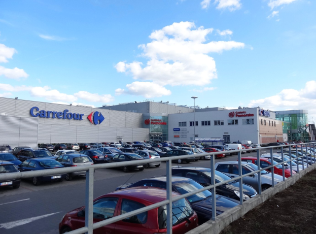 Großes Autohaus mit zahlreichen parkenden Fahrzeugen, umgeben von Gebäuden, Straßeninfrastruktur und Fahrzeugen auf der Straße unter einem bewölkten Himmel.