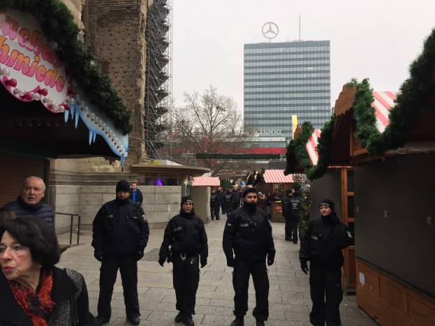 Polizisten in schwarzen Uniformen stehen vor einem Berliner Weihnachtsmarkt, der mit festlichen Dekorationen geschmückt ist, mit Gebäuden, Bäumen und einem klaren blauen Himmel im Hintergrund.