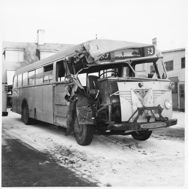Ein Schwarz-Weiß-Foto eines beschädigten Busses am Straßenrand mit eingedellter Front und zersplitterten Fenstern und Gebäuden im Hintergrund.