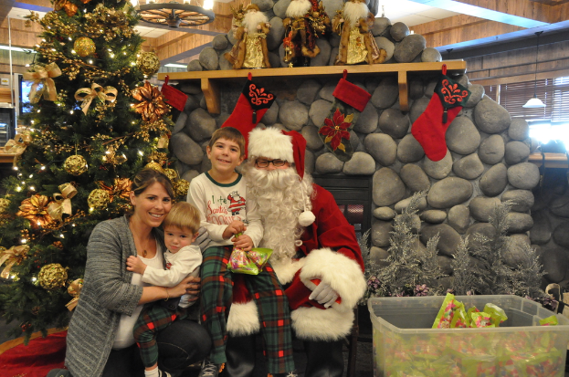 Eine Frau, Kinder und ein Mann als Santa Claus mit Brille und Handschuhen stehen neben einem geschmückten Weihnachtsbaum, einer Wand mit Felsen, Spielsachen, Dekorationen, einem Korb mit Gegenständen und Lichtern im Hintergrund.