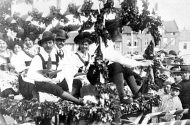 Eine Gruppe von Menschen in traditioneller bayrischer Tracht, einige mit Musikinstrumenten, fährt auf einem mit Blättern geschmückten Wagen in einem Umzug, mit Gebäuden und einem klaren Himmel im Hintergrund, dargestellt in Schwarz-Weiß.