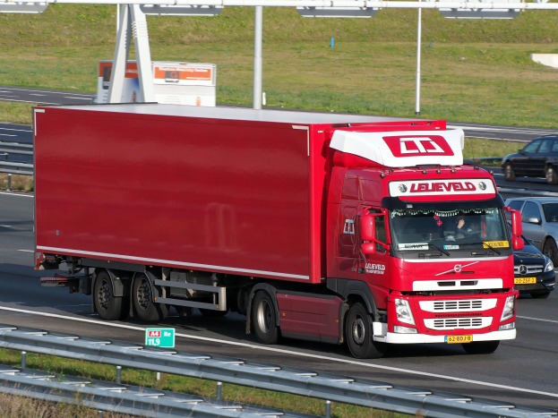 Ein roter Sattelschlepper auf einer Autobahn neben einem schwarzen Auto, mit Gras und einer Begrenzung neben der Straße und Schildern mit Text im Hintergrund.