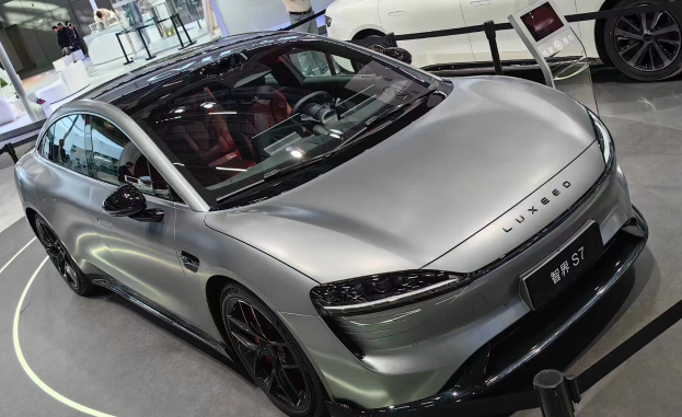 Ein silberner Porsche Taycan auf einer Automesse, umgeben von Menschen, Absperrpoller mit Bändern, einem Display, einer Säule, einer Wand und einer Decke mit Deckenleuchten.