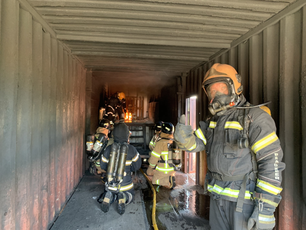Feuerwehrleute in Schutzausrüstung innerhalb eines Containers, einer mit einem Rohr haltend, mit einem Feuerl├Âscher im Hintergrund sichtbar.