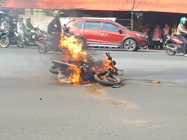 Ein Auto brennt auf der Straße, umgeben von mehreren Helikopter-Leuten und einem Schuppen und Gebäude im Hintergrund.
