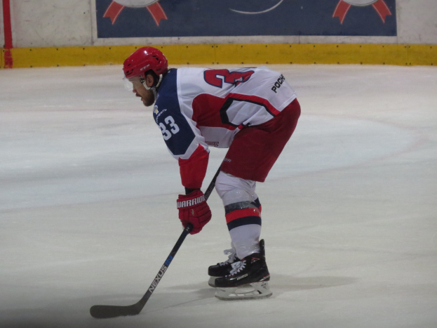 Hockey player on ice in full gear, holding a stick, with a banner featuring text and logos in the background.