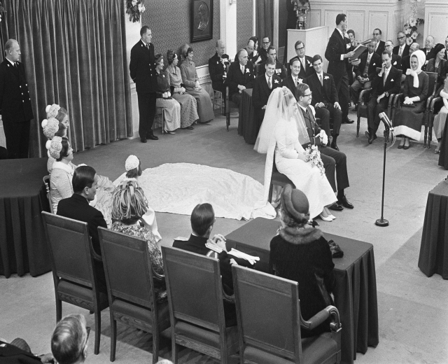 Schwarzes und weißes Foto einer Hochzeitszeremonie mit Menschen, die sitzen und stehen, einem Mann mit Mikrofon, einer Frau mit Blumen, einem Podium mit Mikrofon, Blumenschmuck, einem Vorhang, einem gerahmten Foto an der Wand und einer Tür im Hintergrund.