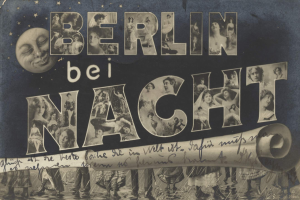 Schwarzes und weißes Foto einer Gruppe von Menschen, die vor einem "Berlin Bei Nacht"-Schild lachen und strahlen, mit einem Mond im Hintergrund, das Schild mit Sternen und Streifen geschmückt.