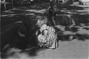 Ein Schwarz-Weiß-Foto von drei Kindern, die im Sand spielen, mit einem Jungen und einem Mädchen, die neben einem Eimer sitzen, und Menschen auf Stühlen mit einem Wagen rechts daneben.